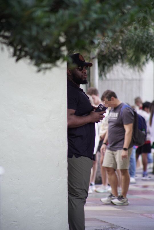 A Black man holding a phone, partly obscured by a wall, watches people in line outdoors.