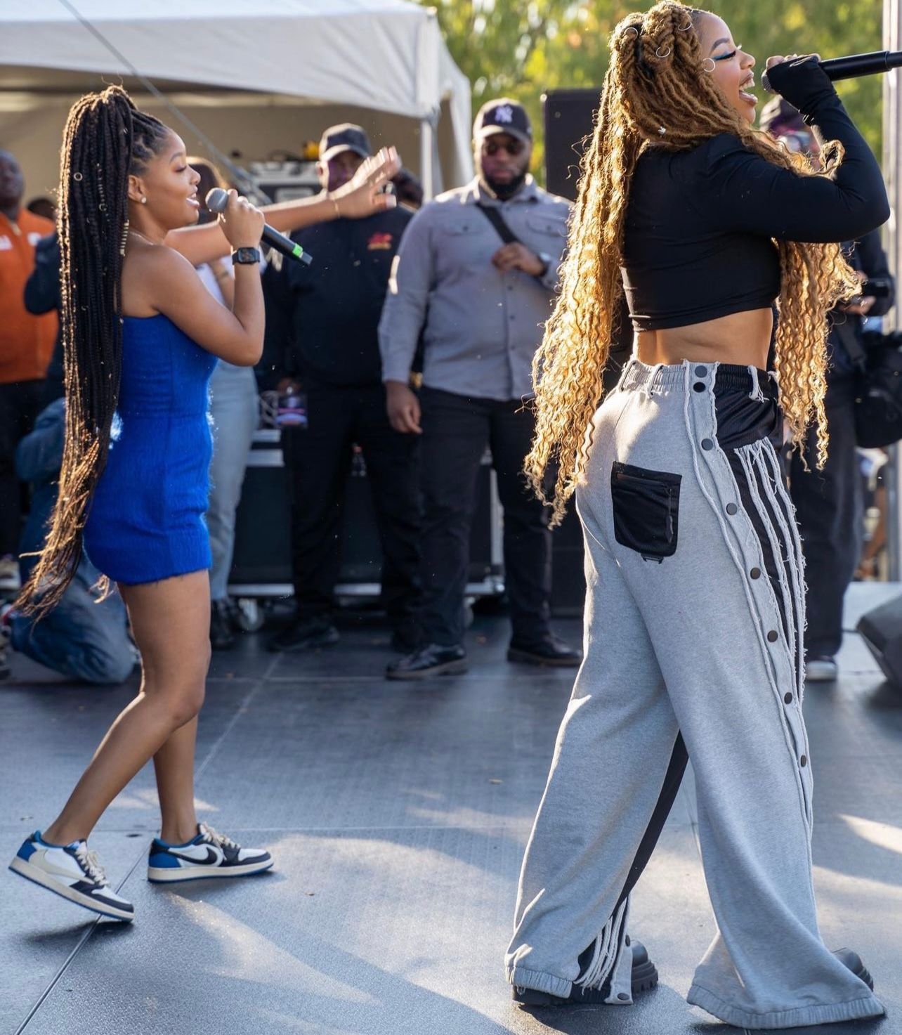 Two women, one in blue dress, the other in gray pants, performing on stage with mics.