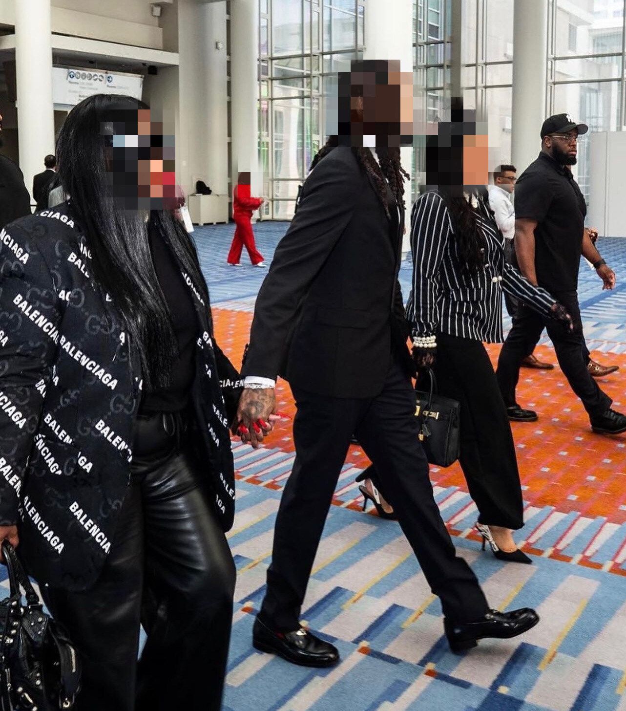 People in formal attire walking in a brightly lit building. A person in a black Balenciaga blazer