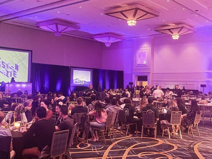 Large event hall with seated guests at round tables, lit by purple lights. Two screens, stage at the front.