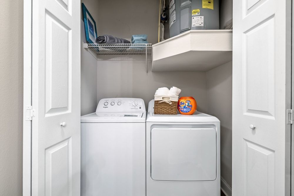 Interior of a neat laundry closet with a white top-loading washing machine and matching dryer. The shelf above the appliances holds detergent and folded towels, with additional storage space and a water heater visible at the back. White doors partially frame the image on both sides, and the walls are painted a neutral color at Marq Perimeter in Atlanta, GA.