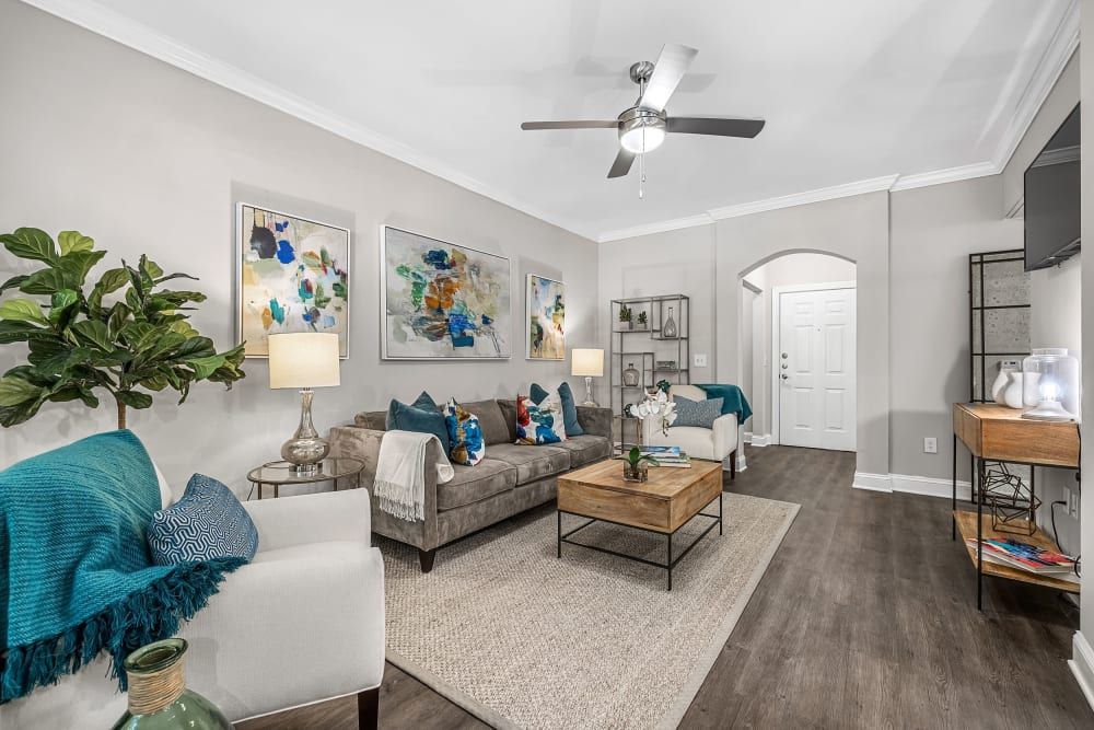 Modern living room interior with neutral tones featuring a gray upholstered sofa with blue throw pillows, matching armchairs, a wooden coffee table atop a beige area rug, and abstract wall art. The room includes a side table with a lamp, a shelf with decorative items, and a ceiling fan. Lush indoor plant adds a touch of greenery, with a visible entrance door in the background at Marq Perimeter in Atlanta, GA.