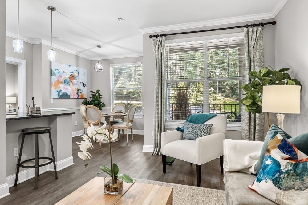 Modern living room interior with a cozy white sofa adorned with colorful pillows, complemented by a simple wood coffee table and elegant white armchairs. A stylish dining set with round table and upholstered chairs is visible in the background, near the kitchen bar with a single stool. The room is illuminated by natural light from a large window with patterned drapes and decorated with a colorful abstract wall painting and indoor plants, creating a harmonious and inviting space at Marq Perimeter in Atlanta, GA.