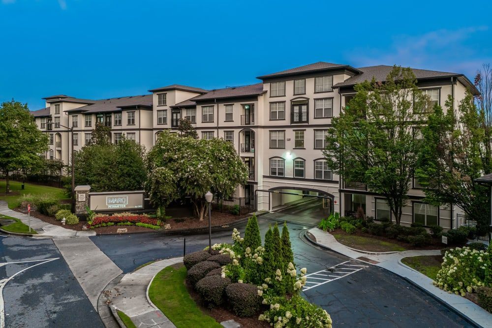 Exterior view of a multi-story residential building with a landscaped entrance and a signboard that reads 'Harrowfield'. The building features a beige facade with dark trim and balconies, surrounded by green trees. The sky is overcast, and there's an empty driveway leading to an underground parking entrance at Marq Perimeter in Atlanta, GA.