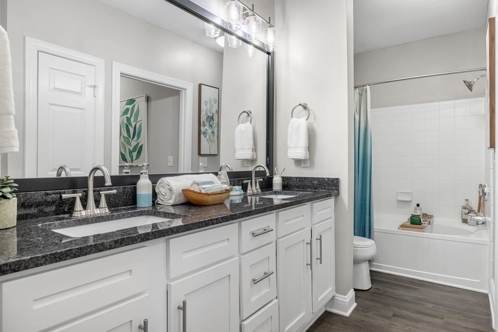 Modern bathroom interior with double sink vanity, mirrors, and white cabinetry. A granite countertop and tiled backsplash complement the white decor. Accessories include towels, soap dispensers, and a blue shower curtain adding a color accent. There is a framed picture on the wall and a toilet is visible beside the bathtub at Marq Perimeter in Atlanta, GA.