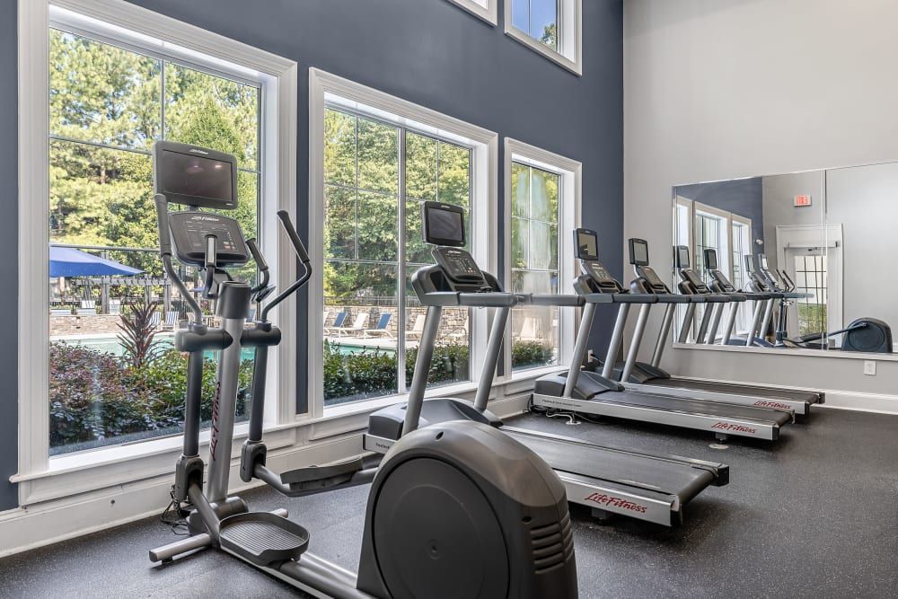 Interior of a well-lit fitness center with multiple cardio machines, including treadmills and an elliptical trainer, facing large windows overlooking a pool area with blue umbrellas and lounge chairs outside at Marq Perimeter in Atlanta, GA.