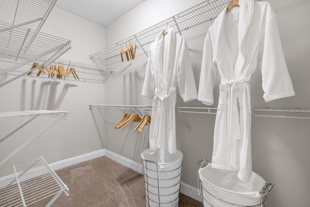 A well-organized closet featuring multiple wire shelving units, with two white bathrobes hanging neatly on wooden hangers, and multiple empty hangers nearby. Two white metal laundry baskets are positioned on the carpeted floor beneath the robes at Marq Perimeter in Atlanta, GA.