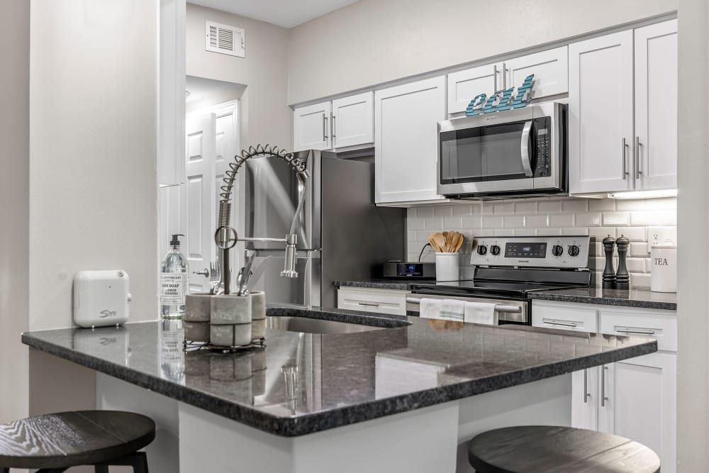 Modern kitchen interior with white cabinetry, stainless steel appliances, and gray granite countertops. Decorative items and kitchen utensils are neatly arranged, including a toaster, a handsoap bottle, and a container holding wooden cooking tools. There's a subway tile backsplash and the natural lighting creates a warm and inviting atmosphere. Two bar stools are positioned under the breakfast bar at Marq Perimeter in Atlanta, GA.