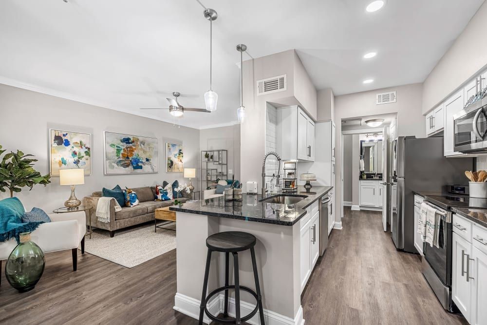 Modern, well-lit open-plan kitchen and living room with neutral color scheme. The kitchen features stainless steel appliances, white cabinetry, and a granite countertop with bar stools, while the living area includes a sofa, armchairs, and wall art with splashes of blue and teal accents at Marq Perimeter in Atlanta, GA.