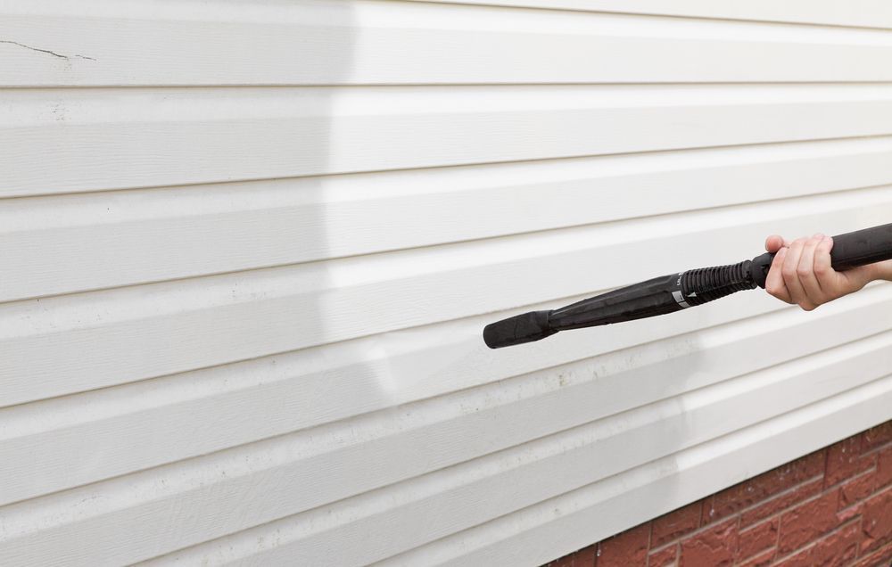 a Person Using a Pressure Washer on White Vinyl Siding Near a Red Brick Foundation — Busy Bees Blasting in Trinity Beach, QLD