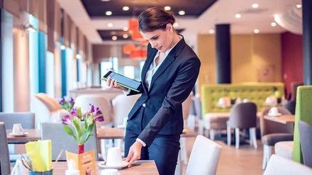 Eine Frau im Anzug steht in einem Restaurant und schaut auf ein Tablet.