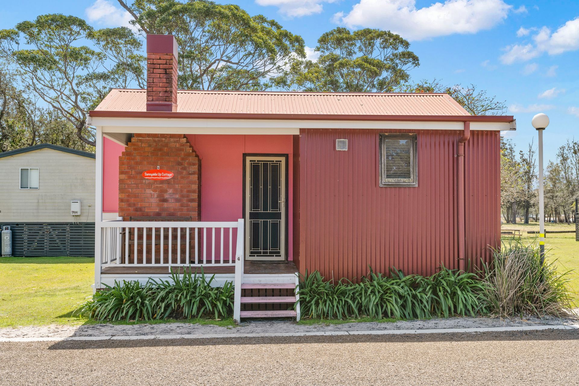 Pink corrugated metal cabin with brick chimney, porch, and small window — SeaChange Parks Milton in Lake Conjola, NSW