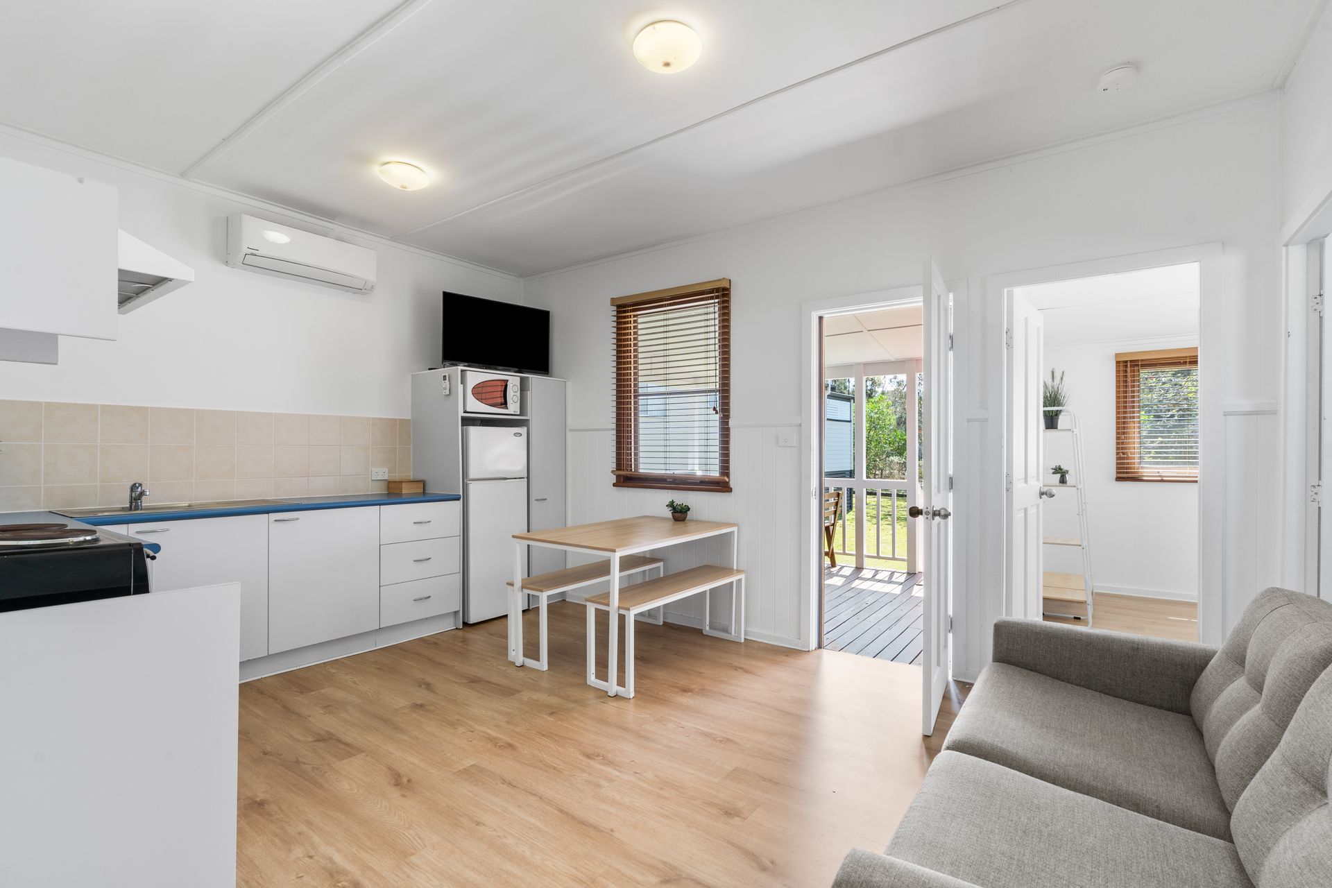 A bright, minimalist studio apartment with a grey sofa, small dining table, kitchenette, and a door leading to a patio. — SeaChange Parks Milton in Lake Conjola, NSW