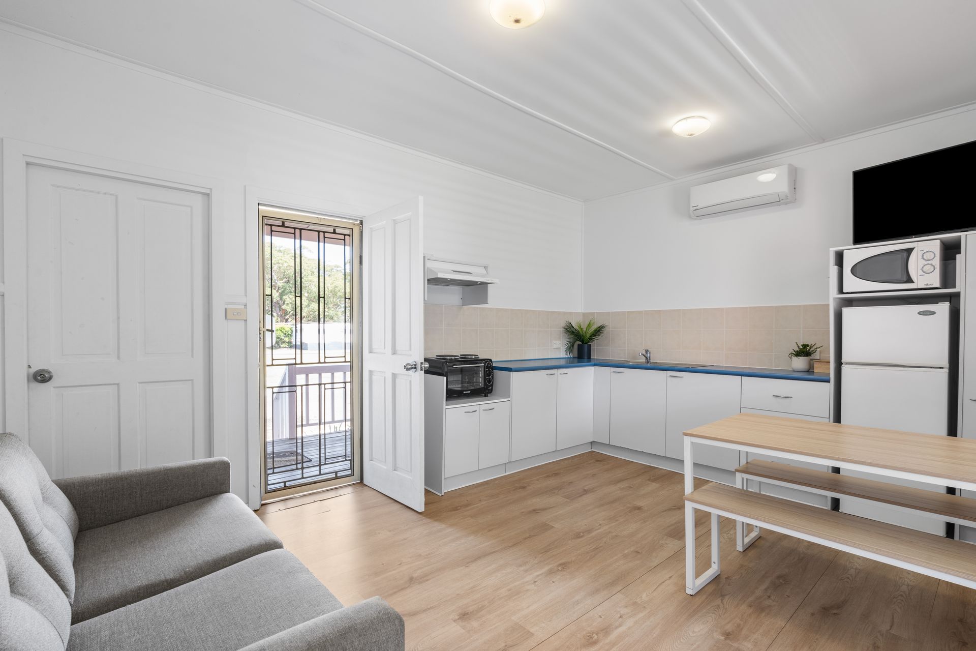 A bright, open-plan studio apartment with a grey sofa, kitchenette, and dining table on light wood flooring. — SeaChange Parks Milton in Lake Conjola, NSW