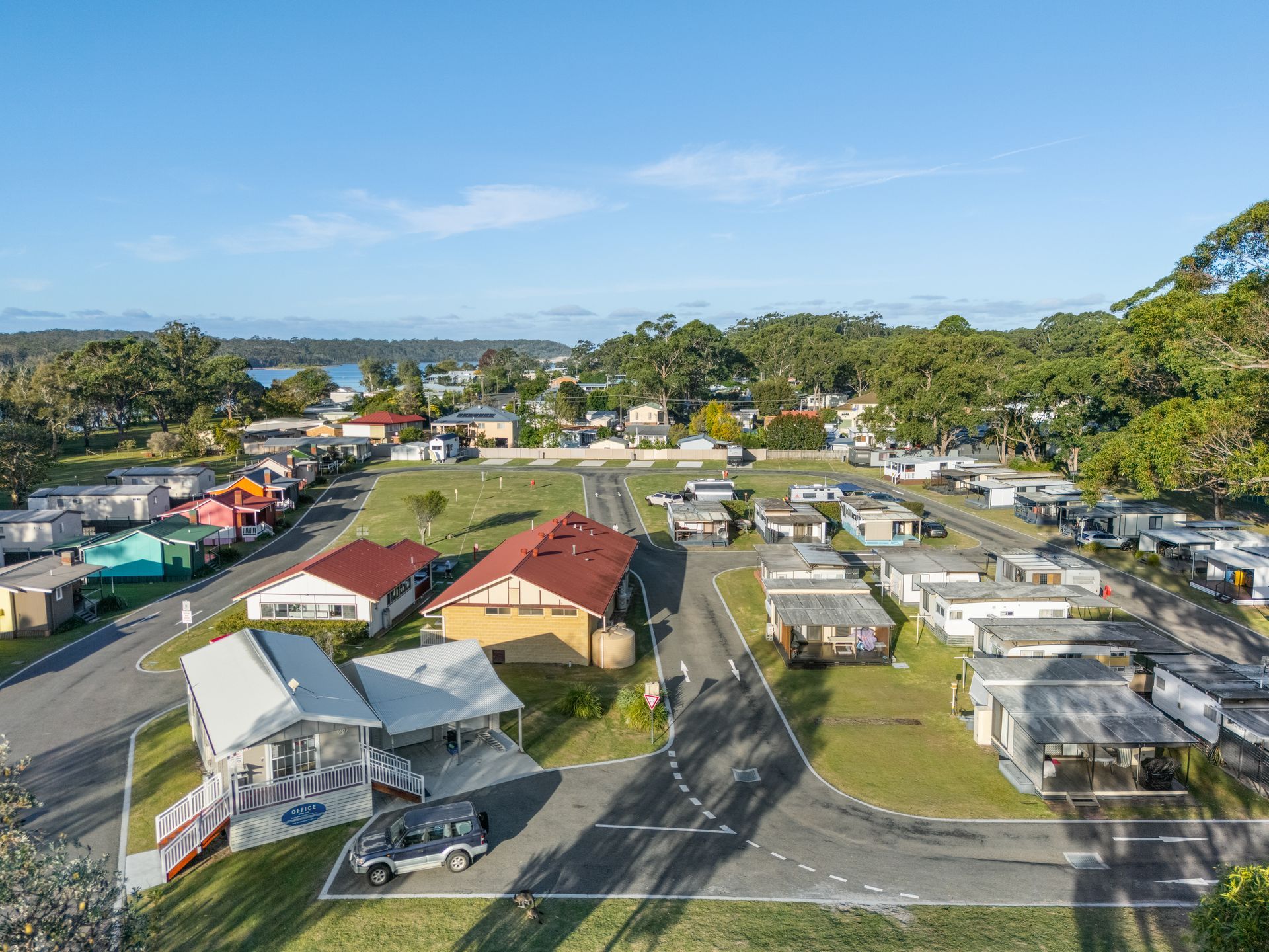 An aerial view of a coastal caravan park with rows of small cabins, mobile homes, and green trees under a clear blue sky. — SeaChange Parks Milton in Lake Conjola, NSW