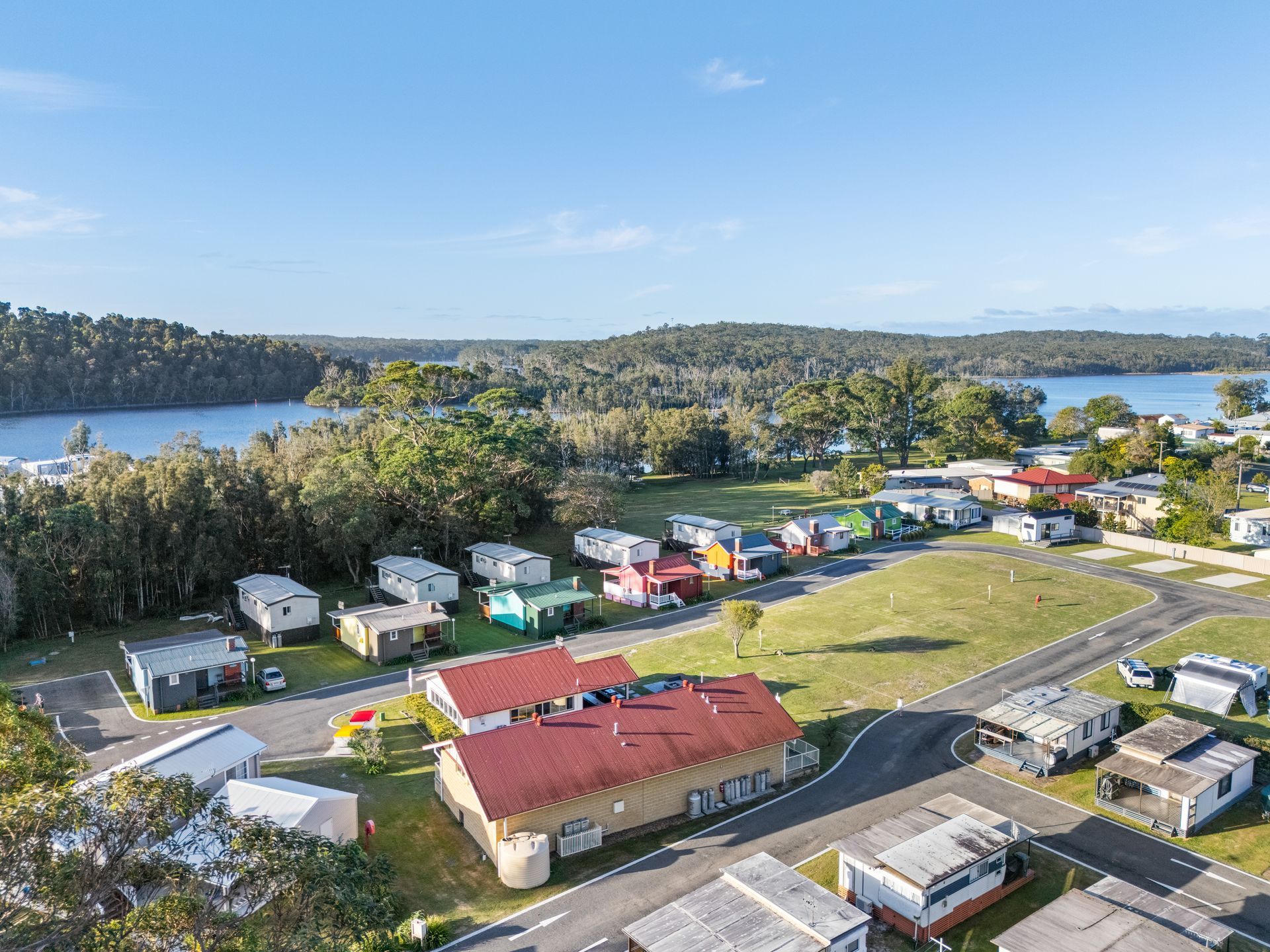 Aerial view of a caravan park with colourful caravans, a building, green grass, trees, and a lake in the background — SeaChange Parks Milton in Milton, NSW