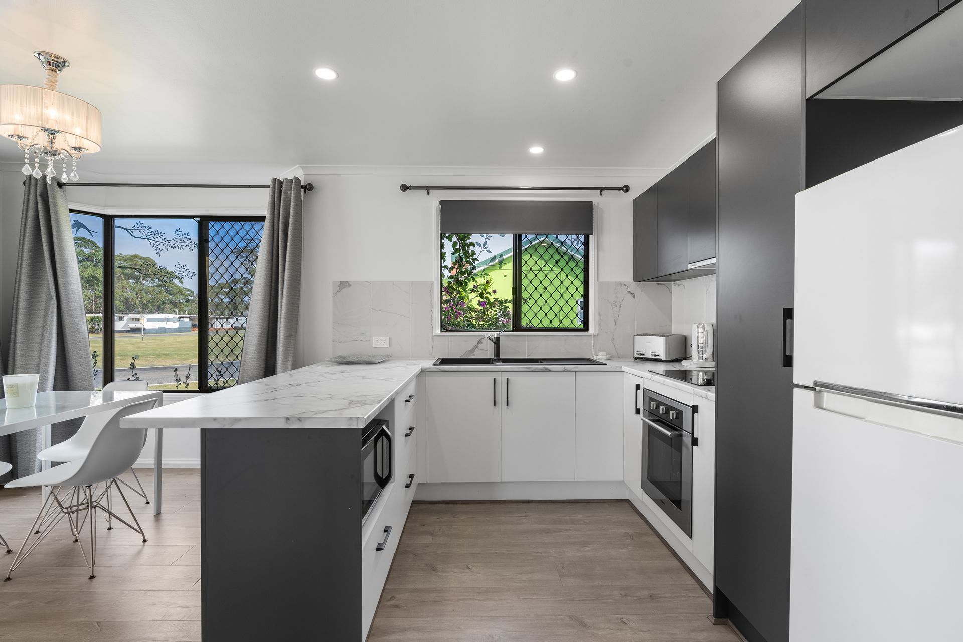 A modern kitchen featuring white cabinets, dark gray accents, marble-look countertops, and a dining area by the window. — SeaChange Parks Milton in Lake Conjola, NSW