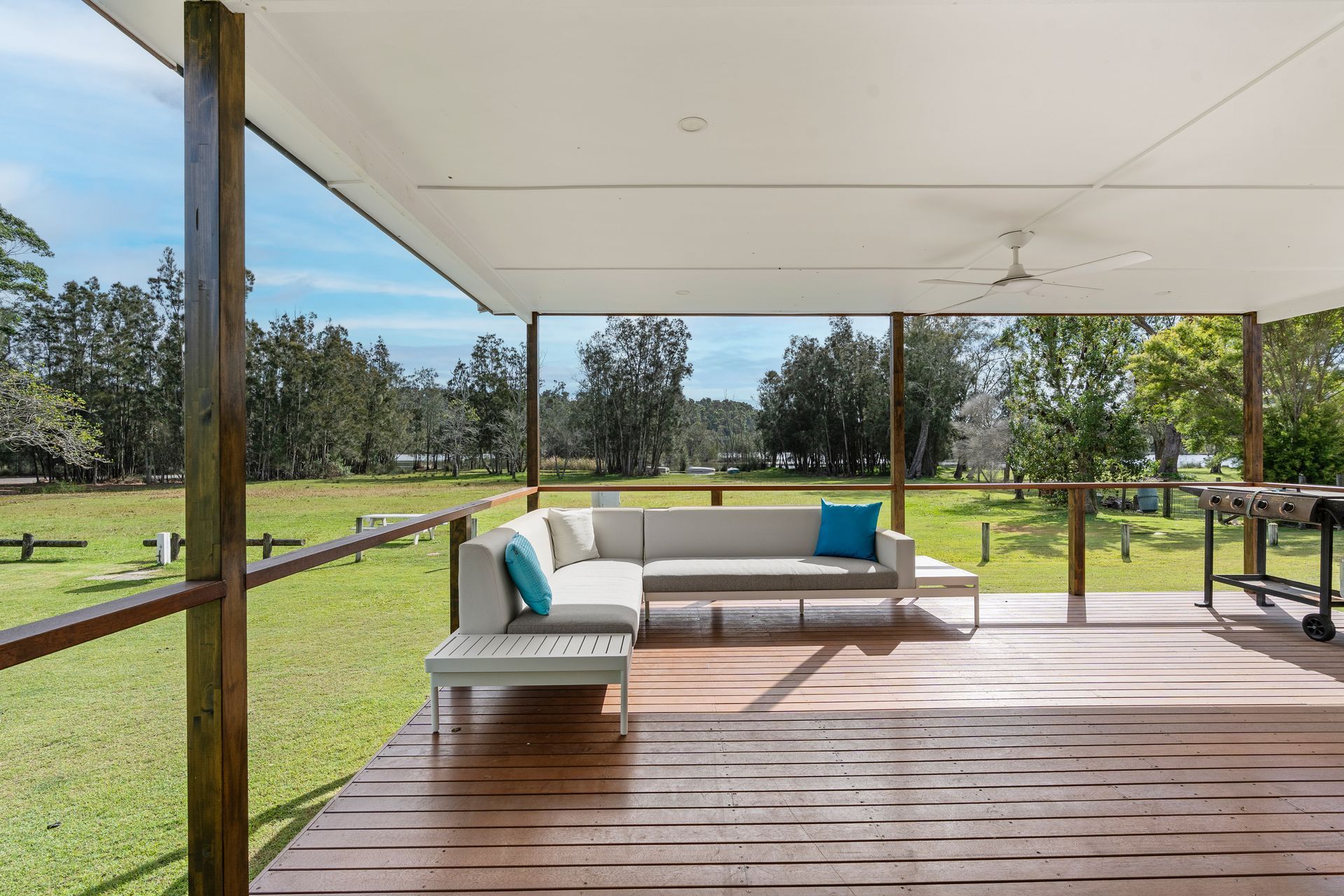 A spacious wooden deck with a white L-shaped outdoor sofa, blue pillows, and a barbecue grill overlooking a grassy field. — SeaChange Parks Milton in Lake Conjola, NSW