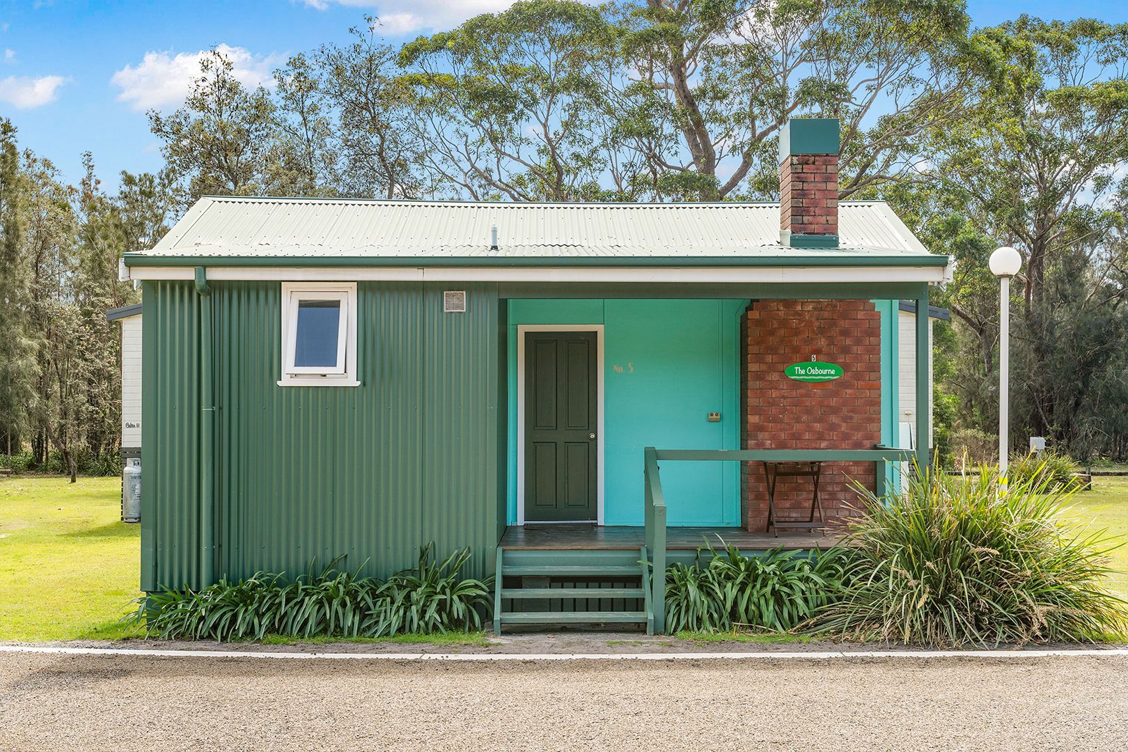 Green and aqua cabin with porch, brick chimney, set on grass near trees — SeaChange Parks Milton in Lake Conjola, NSW