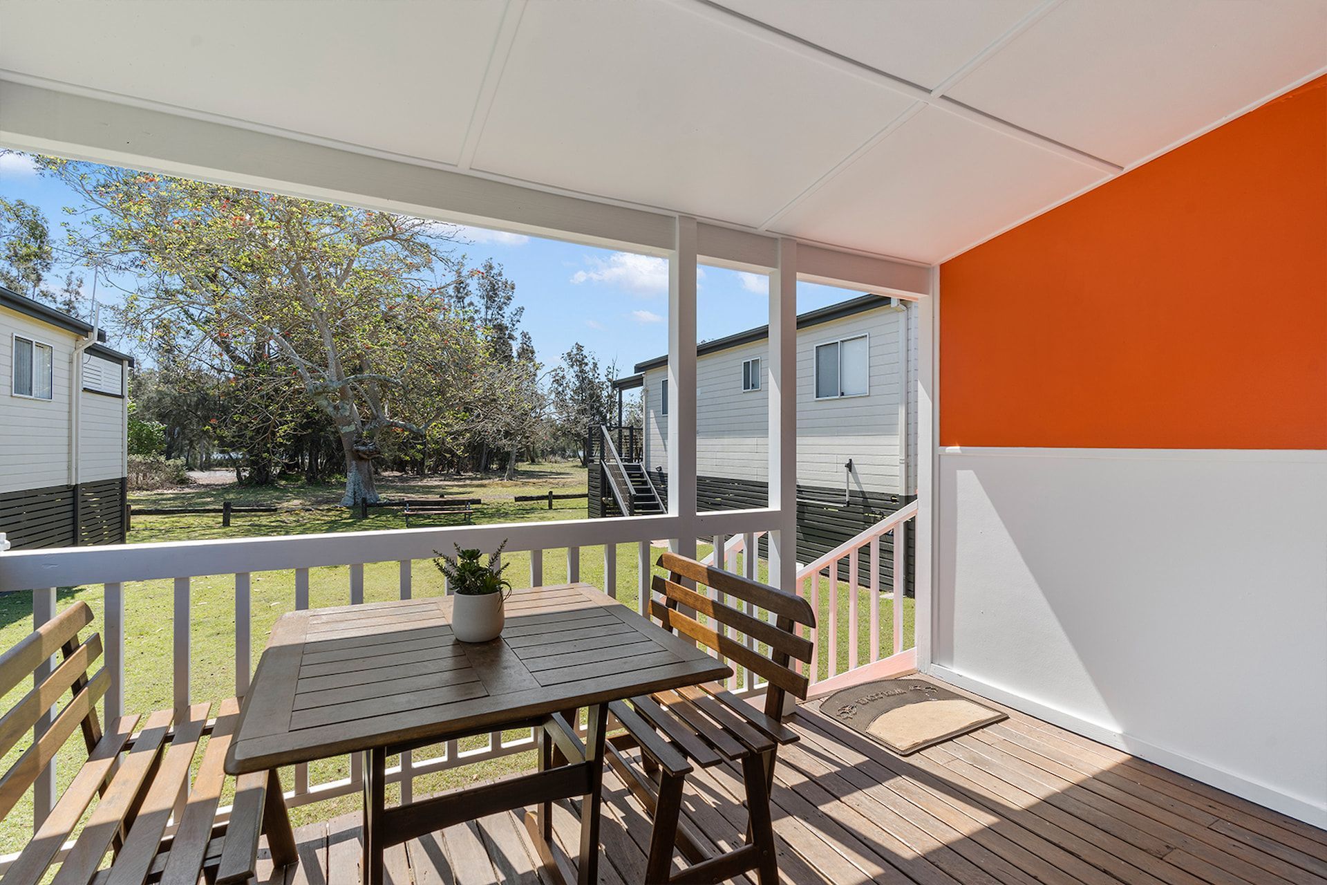 Covered porch with table and chairs overlooking a grassy area and cabins. Orange accent wall — SeaChange Parks Milton in Lake Conjola, NSW