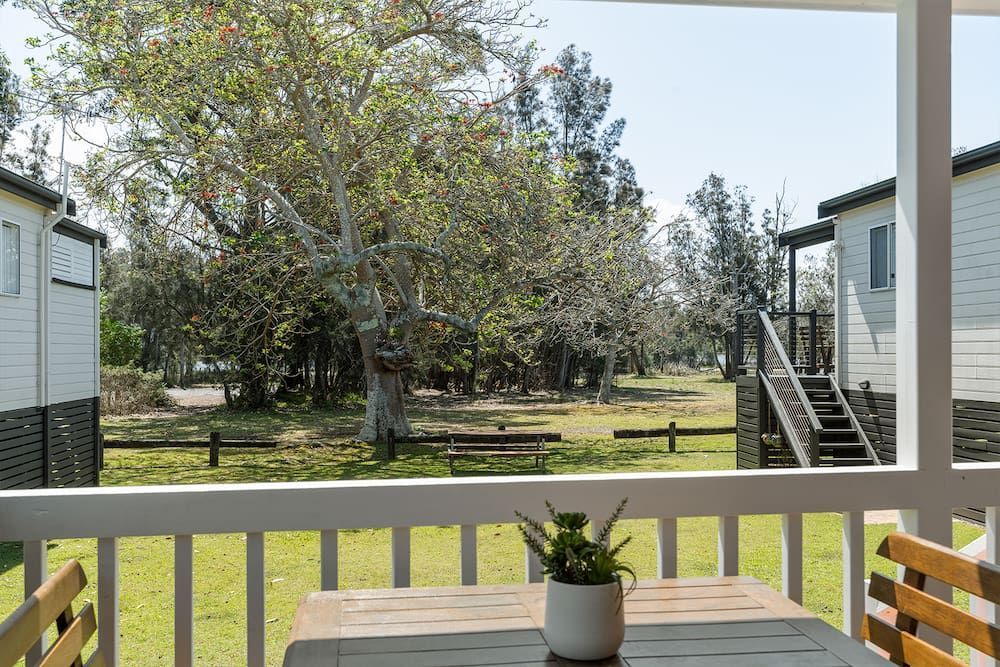 View from a porch with a table and plant, overlooking cabins and trees on a grassy field — SeaChange Parks Milton in Lake Conjola, NSW