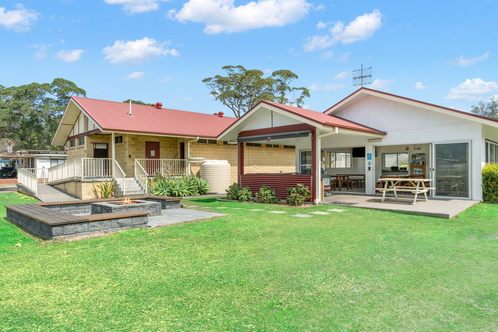 Backyard with a lawn, fire pit, and buildings with red roofs. An outdoor kitchen and seating are visible — SeaChange Parks Milton in Lake Conjola, NSW