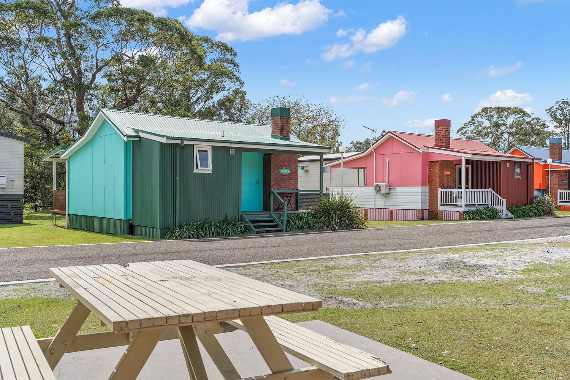 Colourful cabins in a row, with a picnic table in the foreground on a sunny day — SeaChange Parks Milton in Lake Conjola, NSW