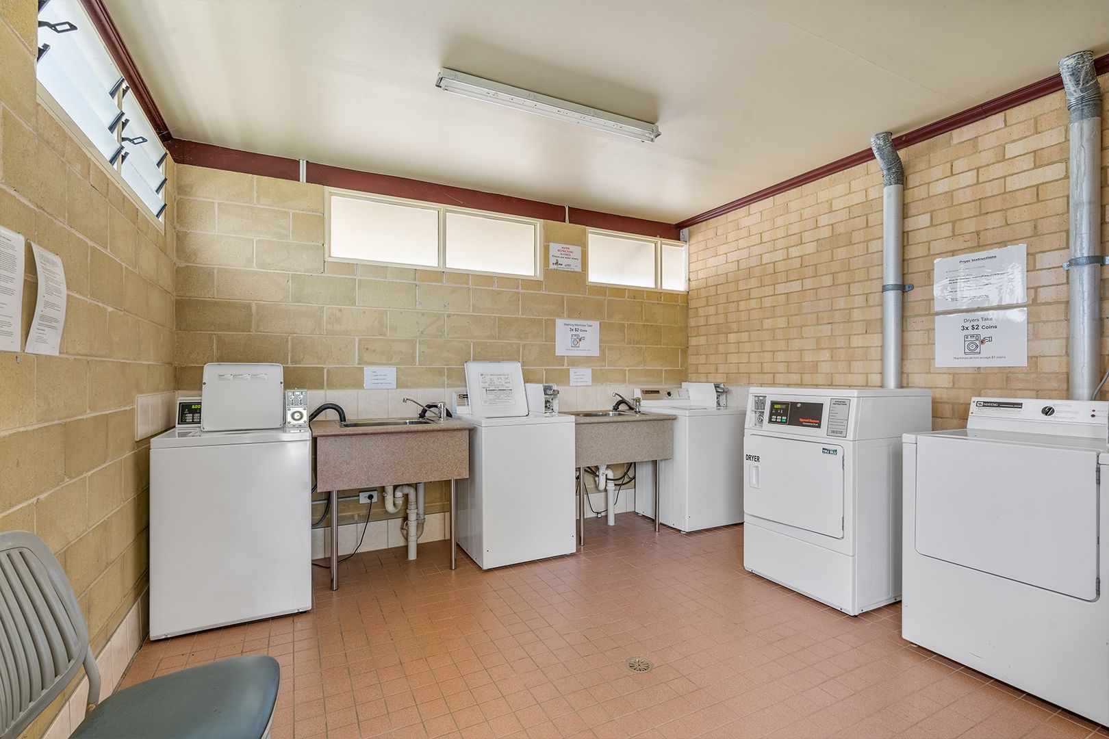Laundry room with several washing machines, sinks, and a chair. Brick and beige walls, with a fluorescent light — SeaChange Parks Milton in Lake Conjola, NSW