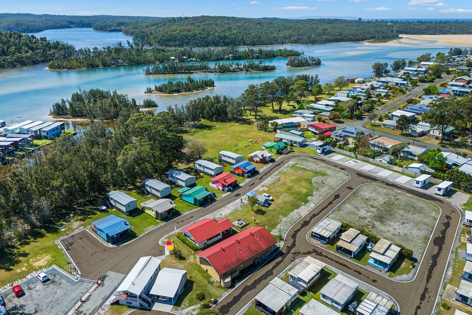 Aerial view: Campground with colourful cabins near a body of water and lush green trees under a blue sky — SeaChange Parks Milton in Lake Conjola, NSW