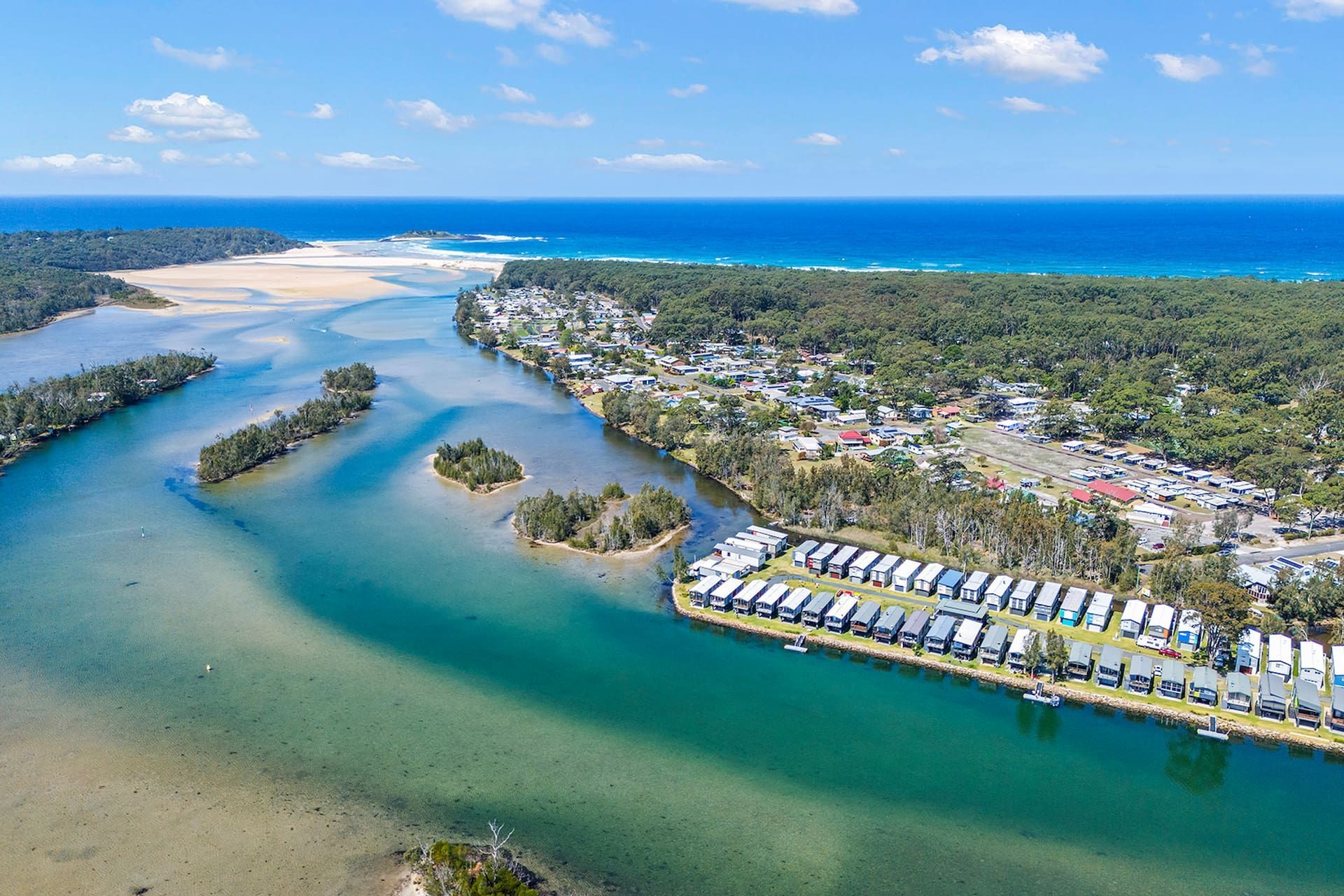 Aerial view: coastal town bordering a river that flows into the ocean, with white buildings and green trees — SeaChange Parks Milton in Lake Conjola, NSW