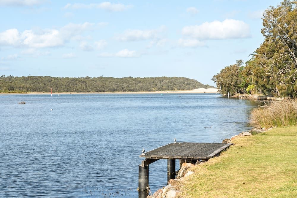 Wooden Dock Extending Into a Wide River, Green Grass on the Bank and Trees — SeaChange Parks Milton in Lake Conjola, NSW