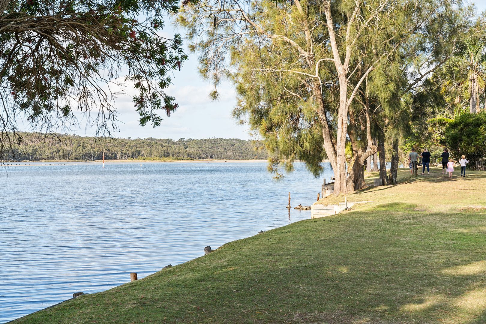 Lakeside park with trees and grass, people walking along the shore on a sunny day — SeaChange Parks Milton in Lake Conjola, NSW