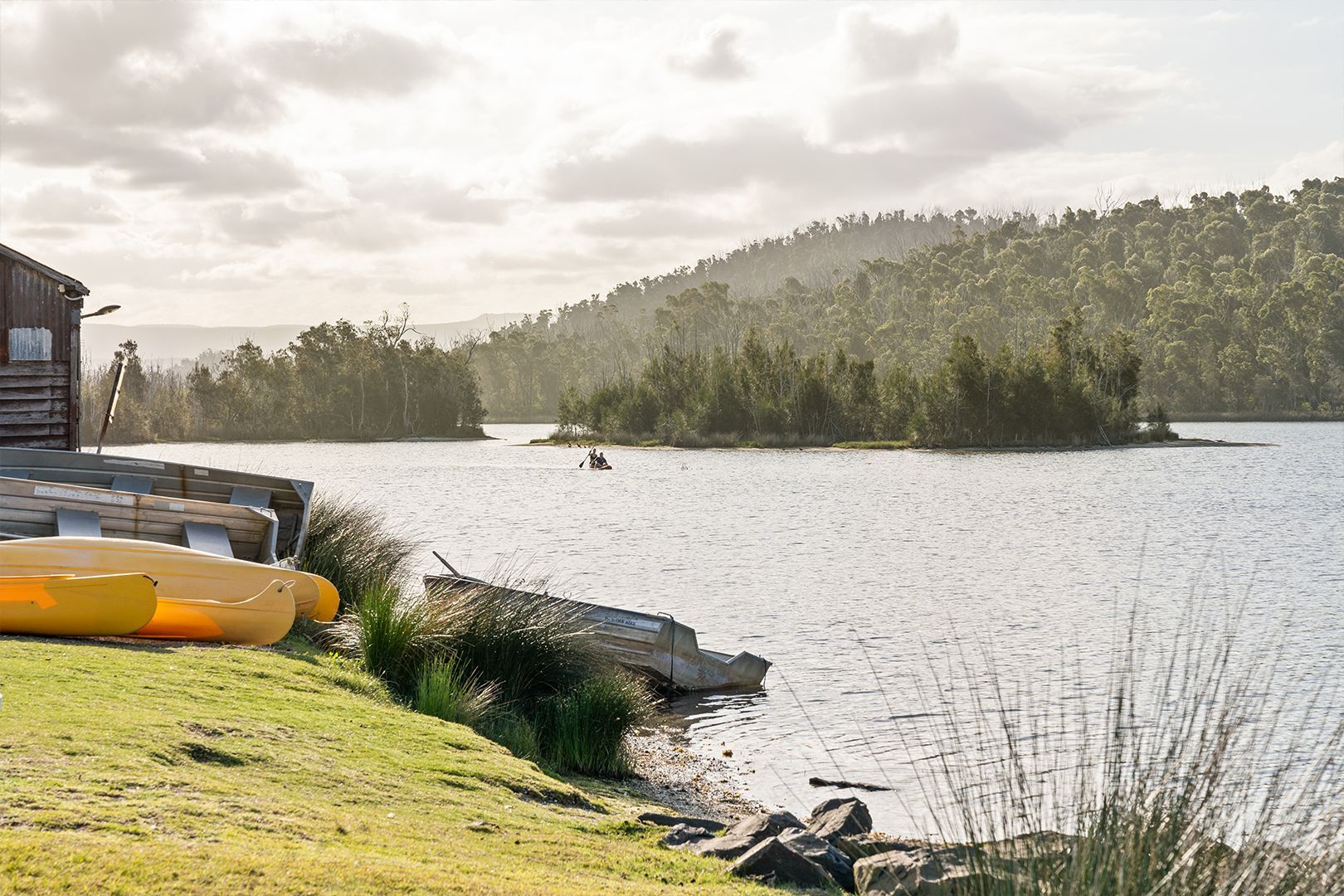 Lakeside scene with small island. Yellow kayaks rest on shore near a wooden building. Mountains in background. — SeaChange Parks Milton in Lake Conjola, NSW