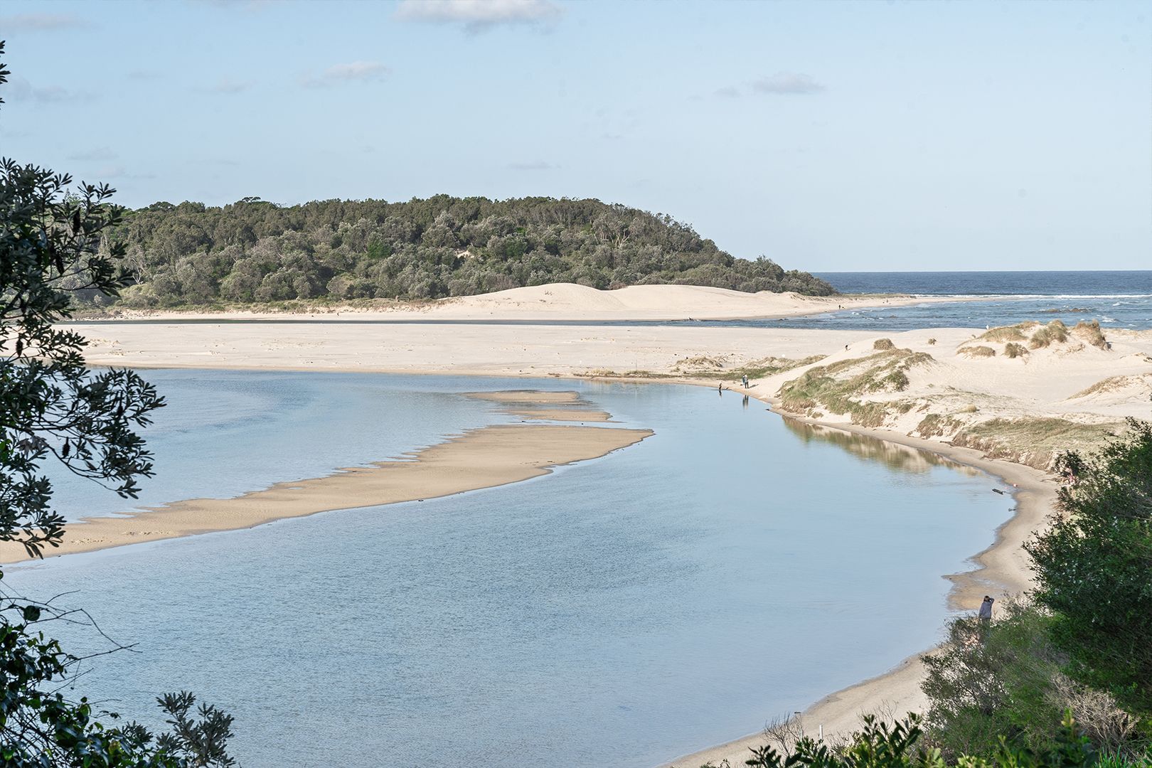 Coastal scene with a sandy beach, river, and forested headland under a blue sky — SeaChange Parks Milton in Lake Conjola, NSW