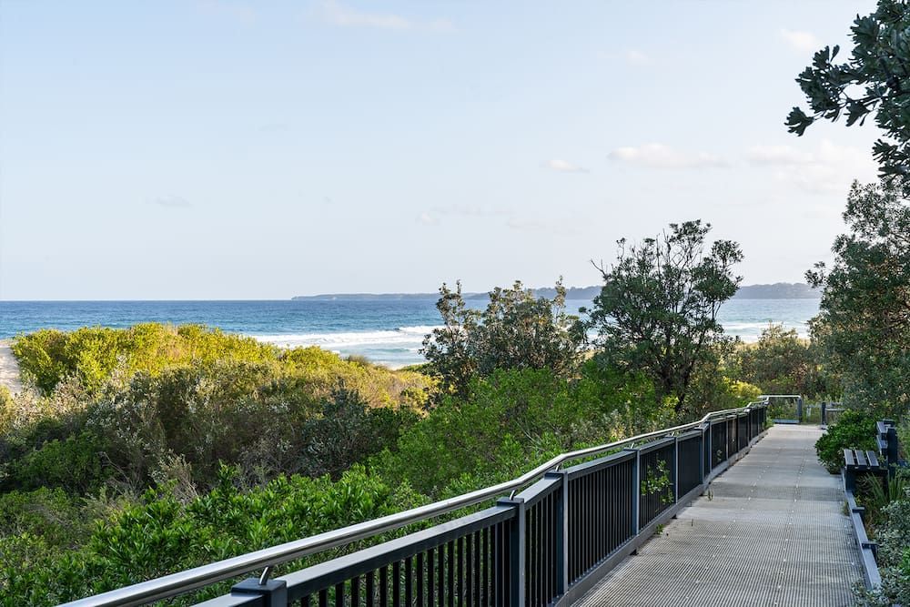 Wooden boardwalk with railings overlooking beach and ocean; green foliage — SeaChange Parks Milton in Lake Conjola, NSW