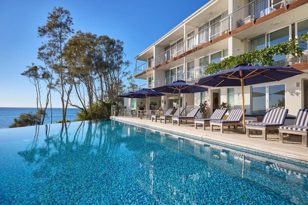 An infinity pool overlooking the ocean, with rows of lounge chairs and blue umbrellas under a resort building — SeaChange Parks Milton in Milton, NSW