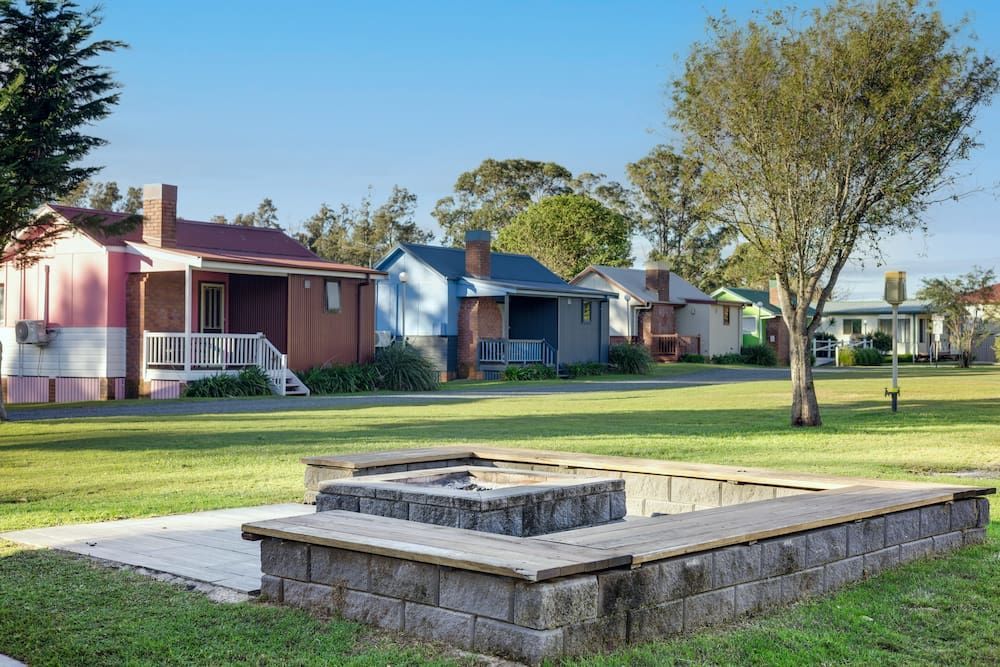 Colourful cottages line a green lawn with a stone fire pit in the foreground under a clear sky — SeaChange Parks Milton in Lake Conjola, NSW