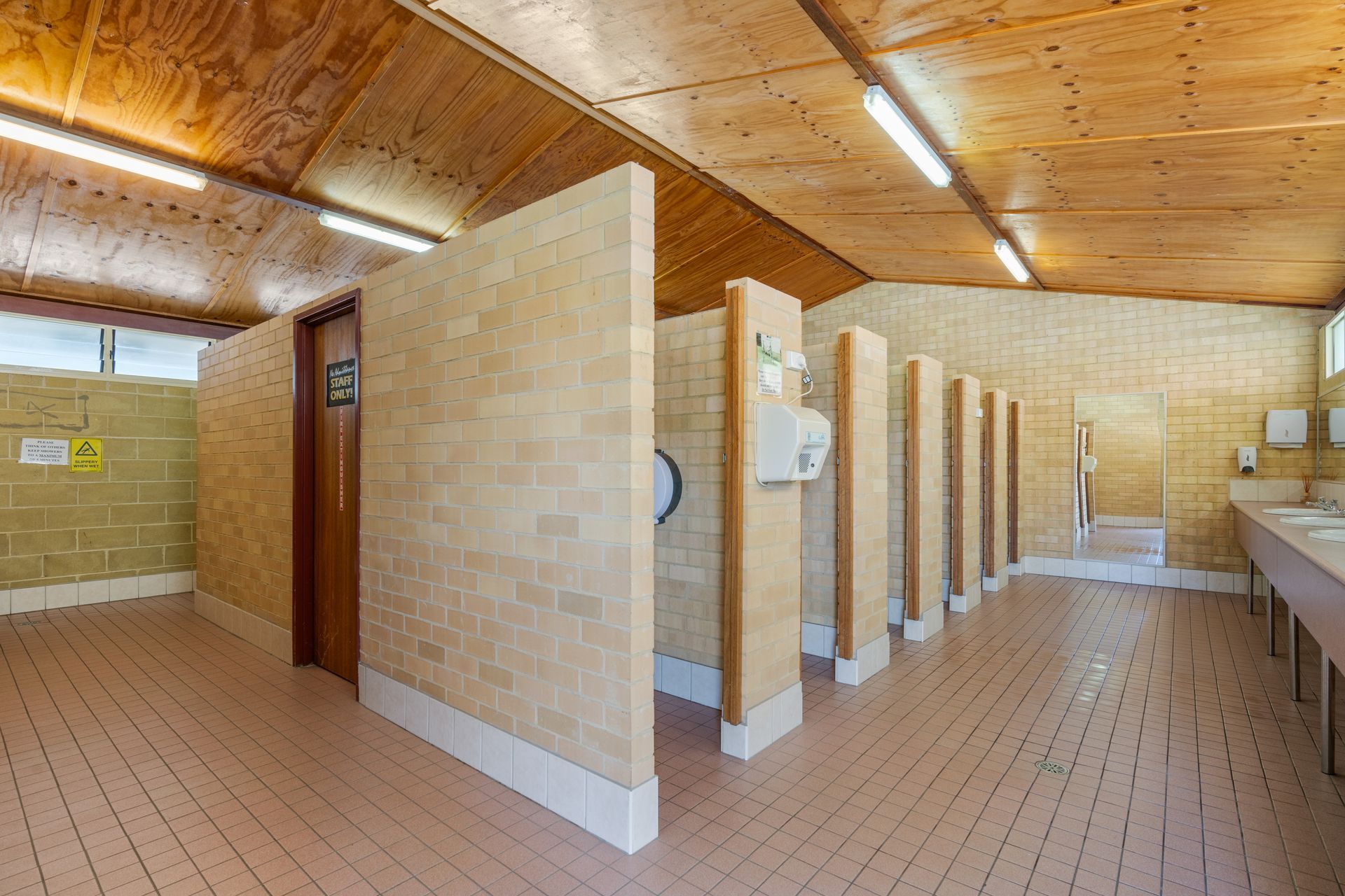 A public restroom with light-coloured brick stall partitions, wooden ceiling, and tiled floor, featuring a row of sinks — SeaChange Parks Milton in Lake Conjola, NSW