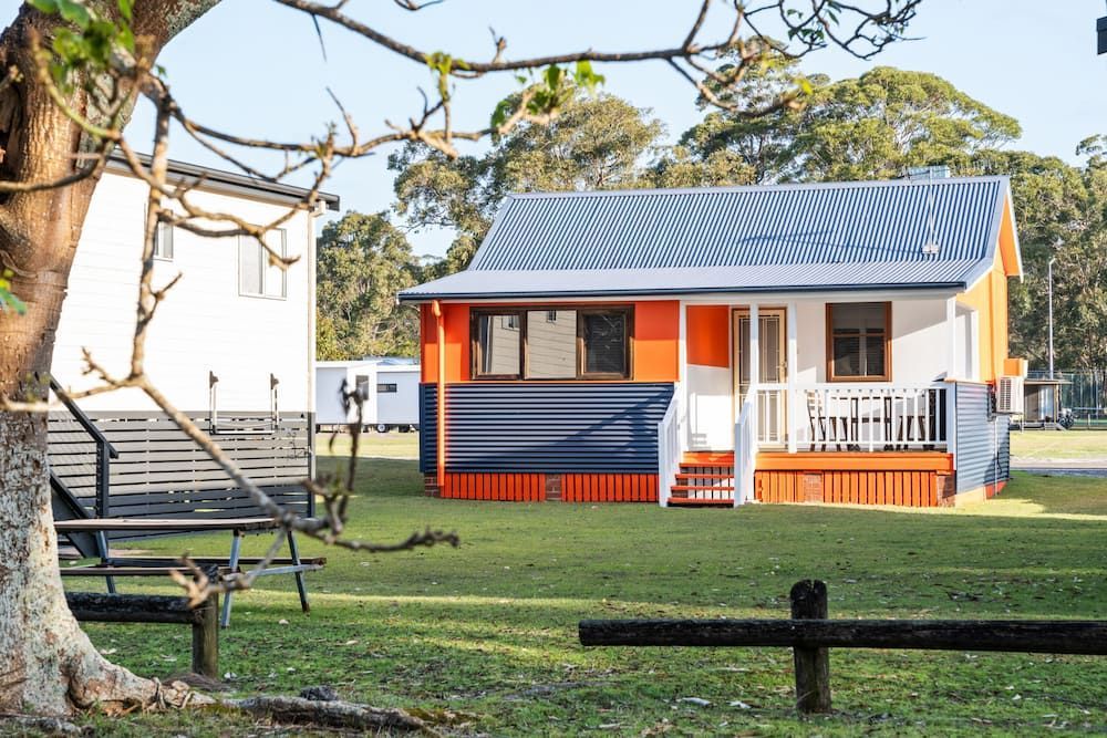 Cottage With Orange and Blue Accents, White Trim, and a Porch, on a Grassy Lawn — SeaChange Parks Milton in Lake Conjola, NSW