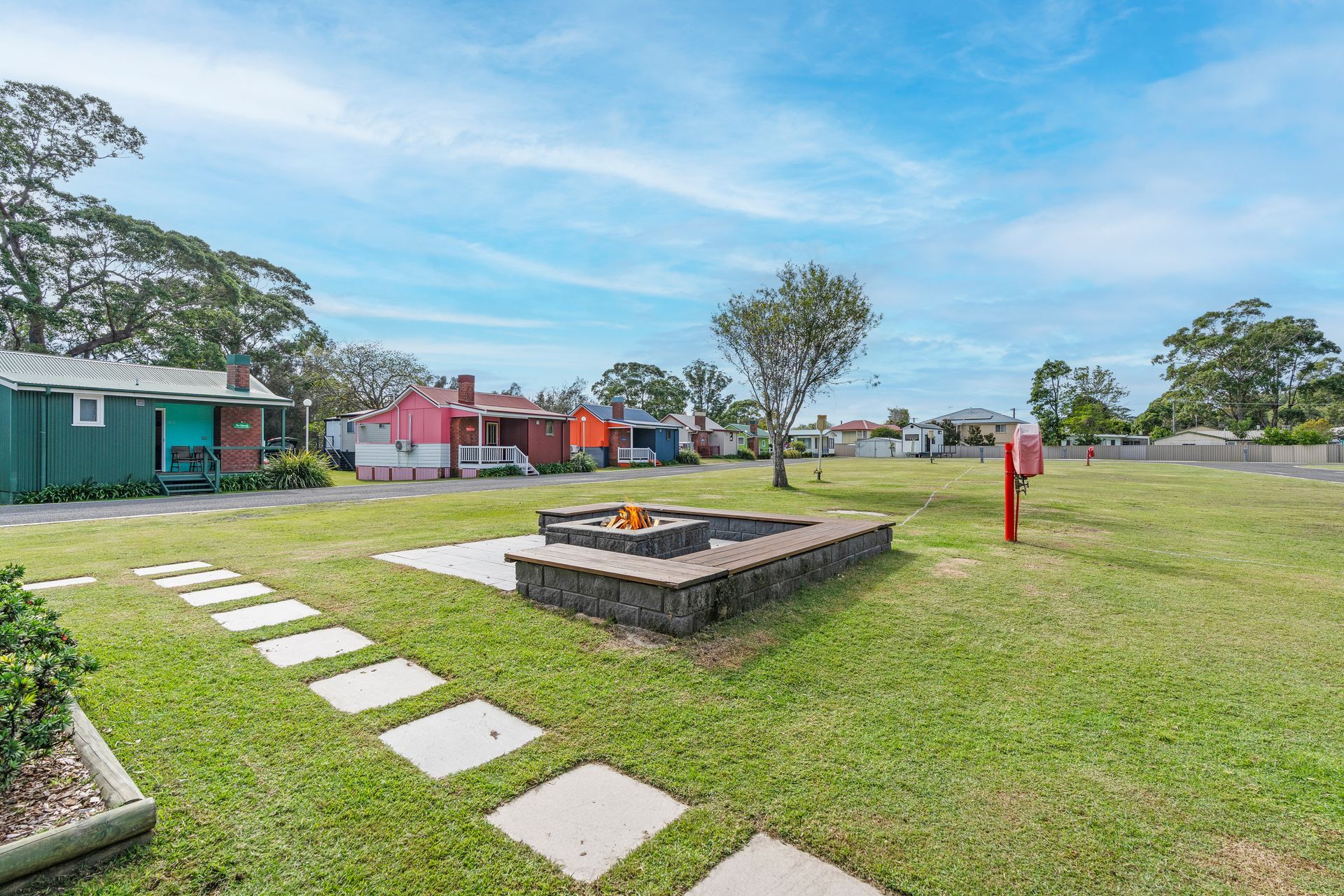 Grassy park with stepping stones, a brick fire pit, colourful cabins — SeaChange Parks Milton in Lake Conjola, NSW