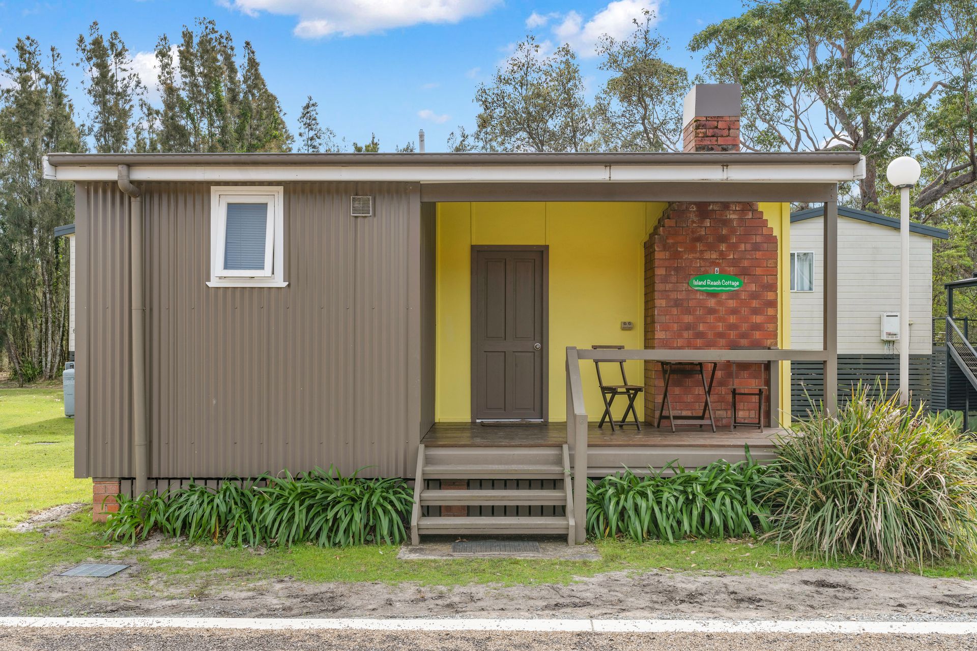 Cabin with yellow door and brown siding, porch with chairs, brick chimney — SeaChange Parks Milton in Lake Conjola, NSW