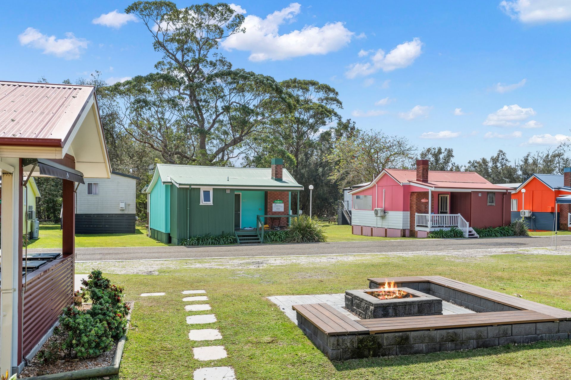 A sunny outdoor area featuring small, colourful cabins with a stone fire pit in the foreground on a grass lawn — SeaChange Parks Milton in Milton, NSW