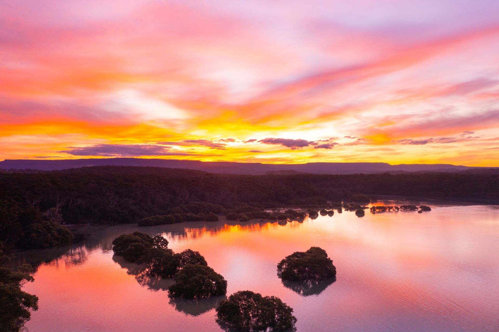 Vibrant sunset with orange and purple clouds reflected in a lake dotted with small islands and trees — SeaChange Parks Milton in Milton, NSW