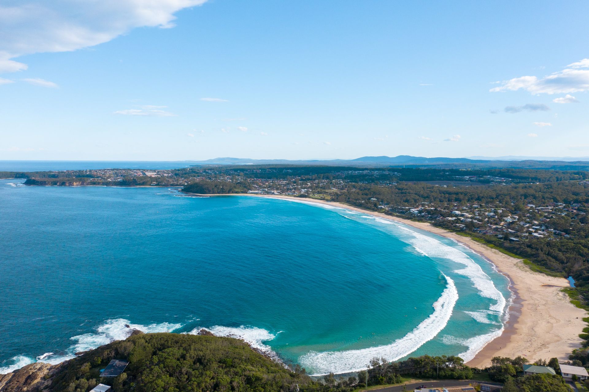 Aerial view of a vibrant turquoise crescent-shaped beach surrounded by dense greenery and a coastal town under a blue sky — SeaChange Parks Milton in Milton, NSW