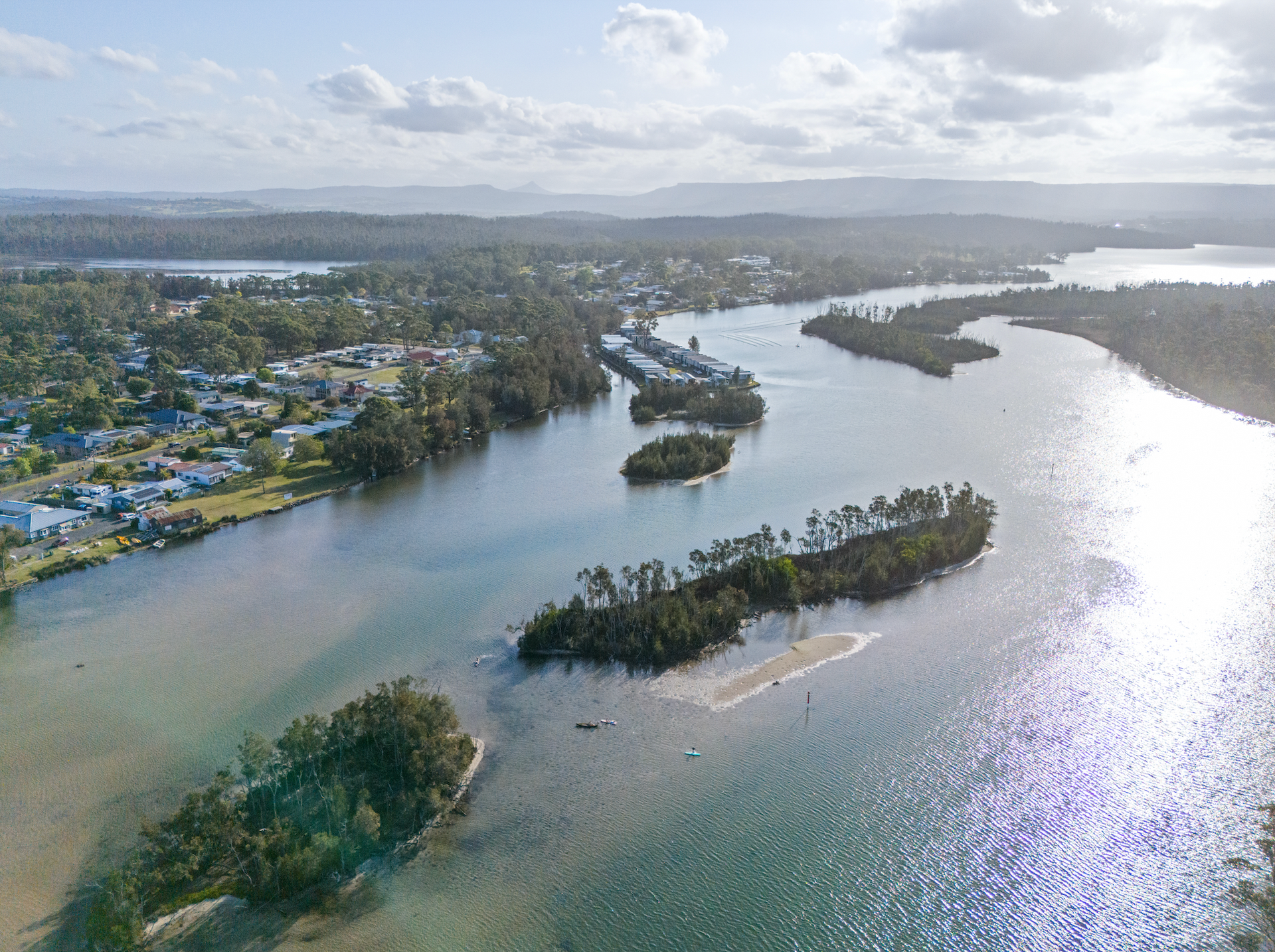Aerial view of a winding river featuring small, tree-covered islands and a residential coastal town in the distance. — SeaChange Parks Milton in Lake Conjola, NSW