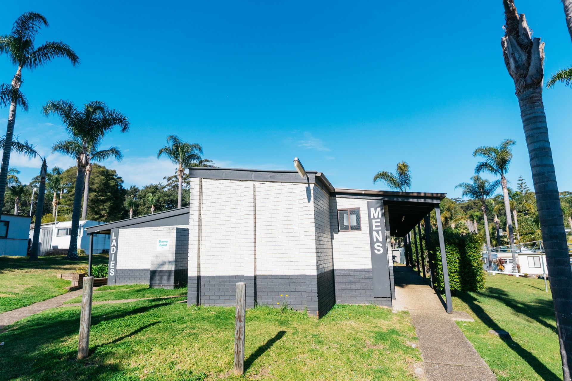 A single-story, utilitarian building with horizontal siding, labeled 'MENS' and 'LADIES', surrounded by palm trees  — SeaChange Parks Milton in Milton, NSW