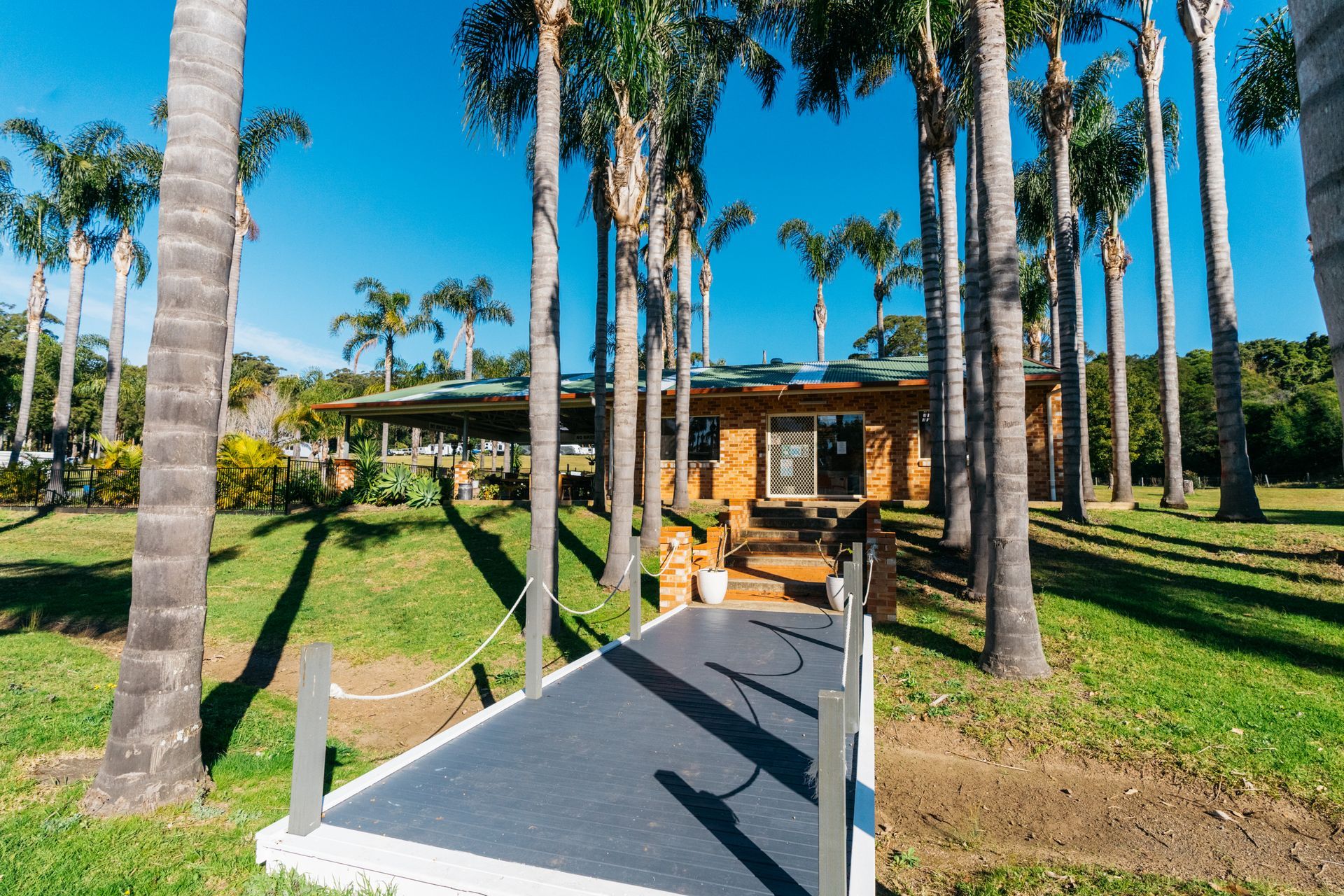 A paved walkway leads to a brick building surrounded by tall palm trees under a bright blue sky  — SeaChange Parks Milton in Milton, NSW