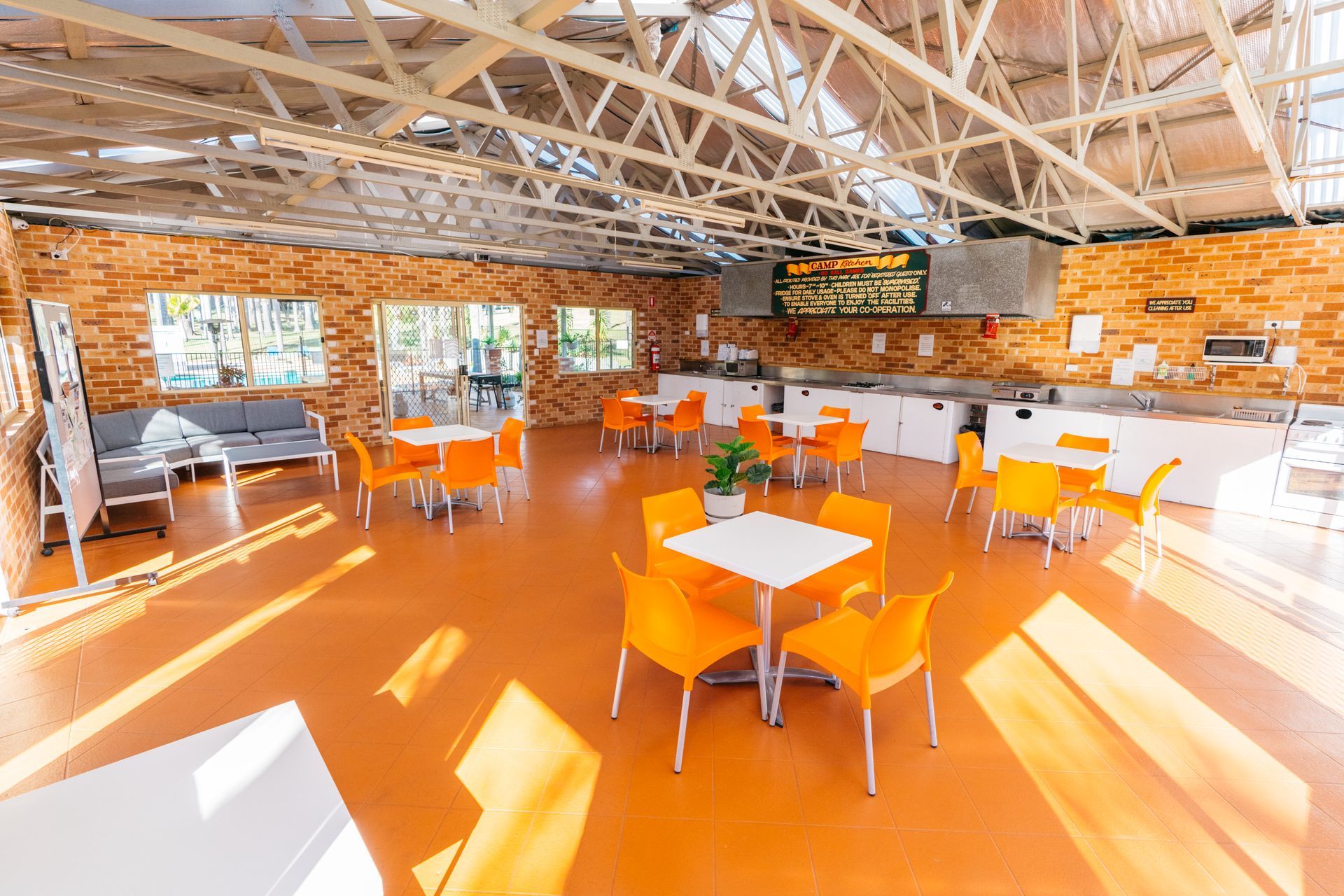 An open-air dining area with orange floors, white tables, and orange chairs under a vaulted metal roof — SeaChange Parks Milton in Milton, NSW