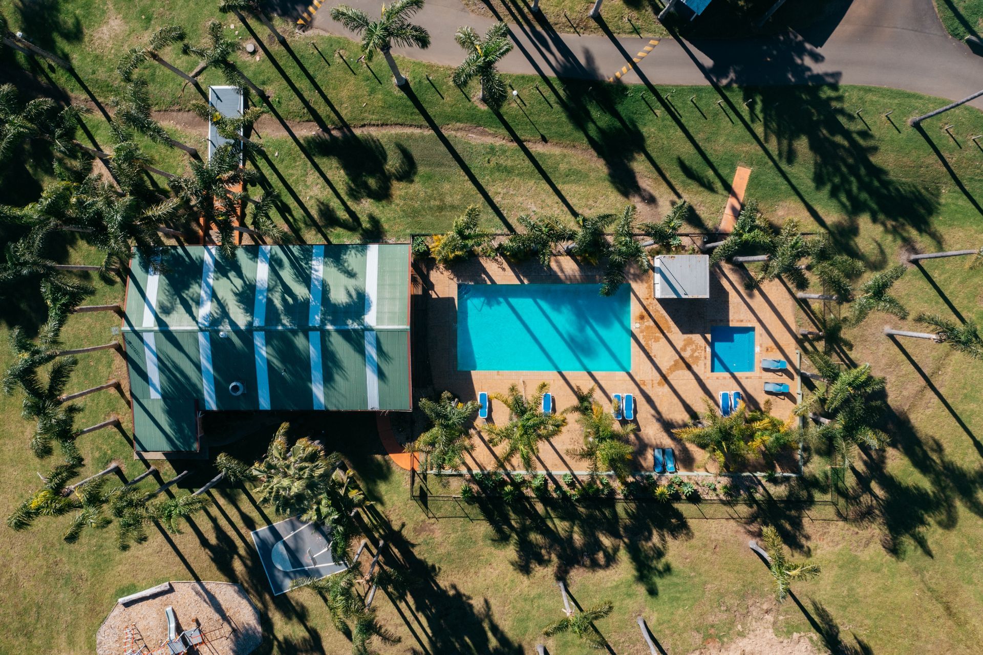 An aerial view of a green-roofed building next to a bright blue swimming pool, surrounded by palm trees on a grassy lawn  — SeaChange Parks Milton in Milton, NSW