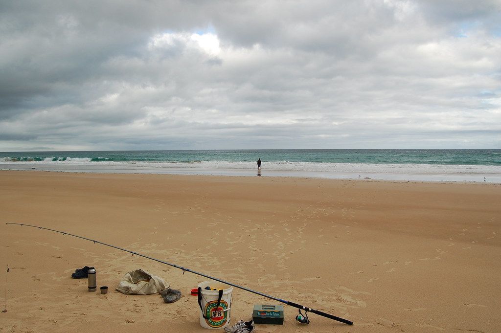 Fishing gear laid out on a sandy beach with a person standing in the ocean surf under a cloudy sky — SeaChange Parks Milton in Milton, NSW